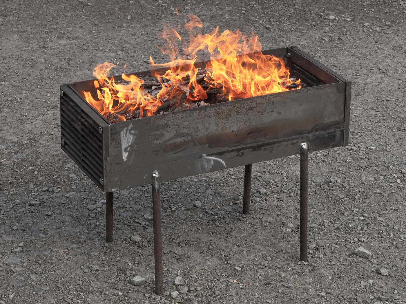 A rectangular steel volume that is open at the top. There are air slots in the head ends. The object stands on 4 thin legs, a fire burns inside the volume.