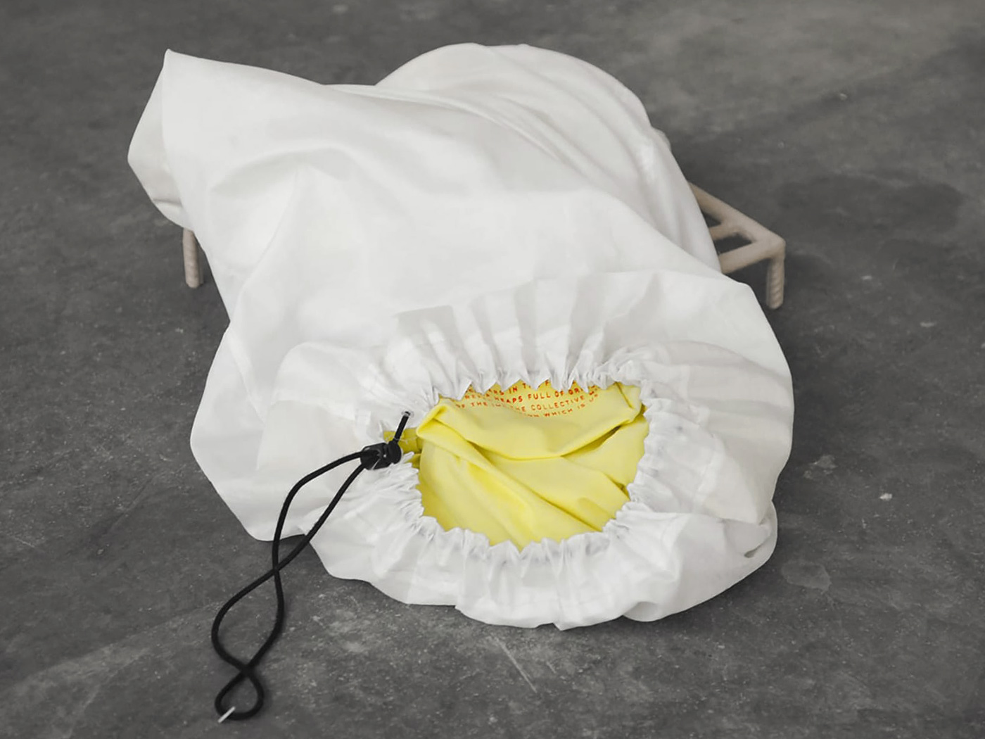 A white laundry bag made of ripstop fabric, the drawstring closure is slightly open. The bag contains a signal yellow item of clothing with red lettering printed on it.