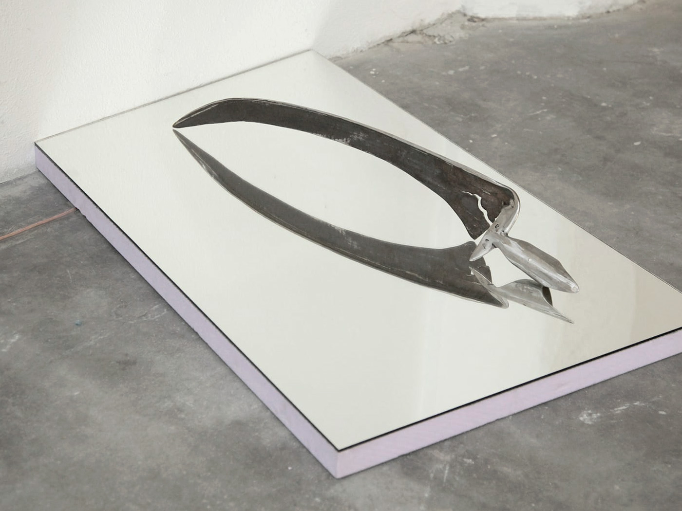 Modified steel scythe on a mirror surface. Under the reflective surface, a base made of lilac-colored foam and a copper cable running out of the picture to the left.