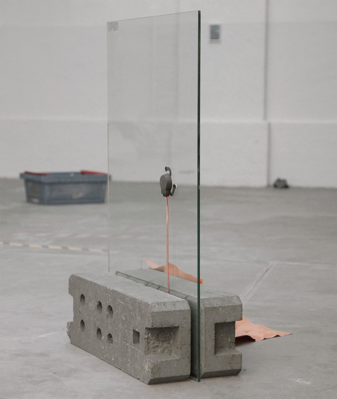 A glass pane in portrait format, to which an audio exciter is attached. The glass pane is held to the floor by two construction fence weights.