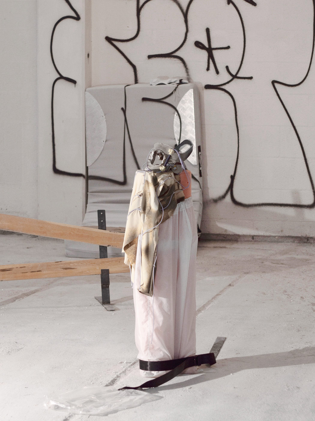 A neck guard on a fabric base. A textile torso hangs down from the base. A rudimentary barrier, a mattress leaning against the wall and graffiti can be seen in the background.