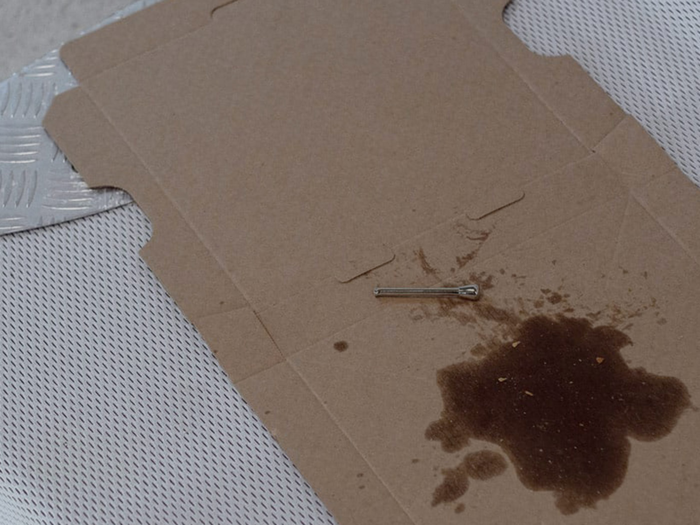 A snorting tube on an unfolded pizza box, which is placed on a mattress. There is a large residue of fat underneath the snorting tube. At the corner of the mattress is an aluminum stair guard.