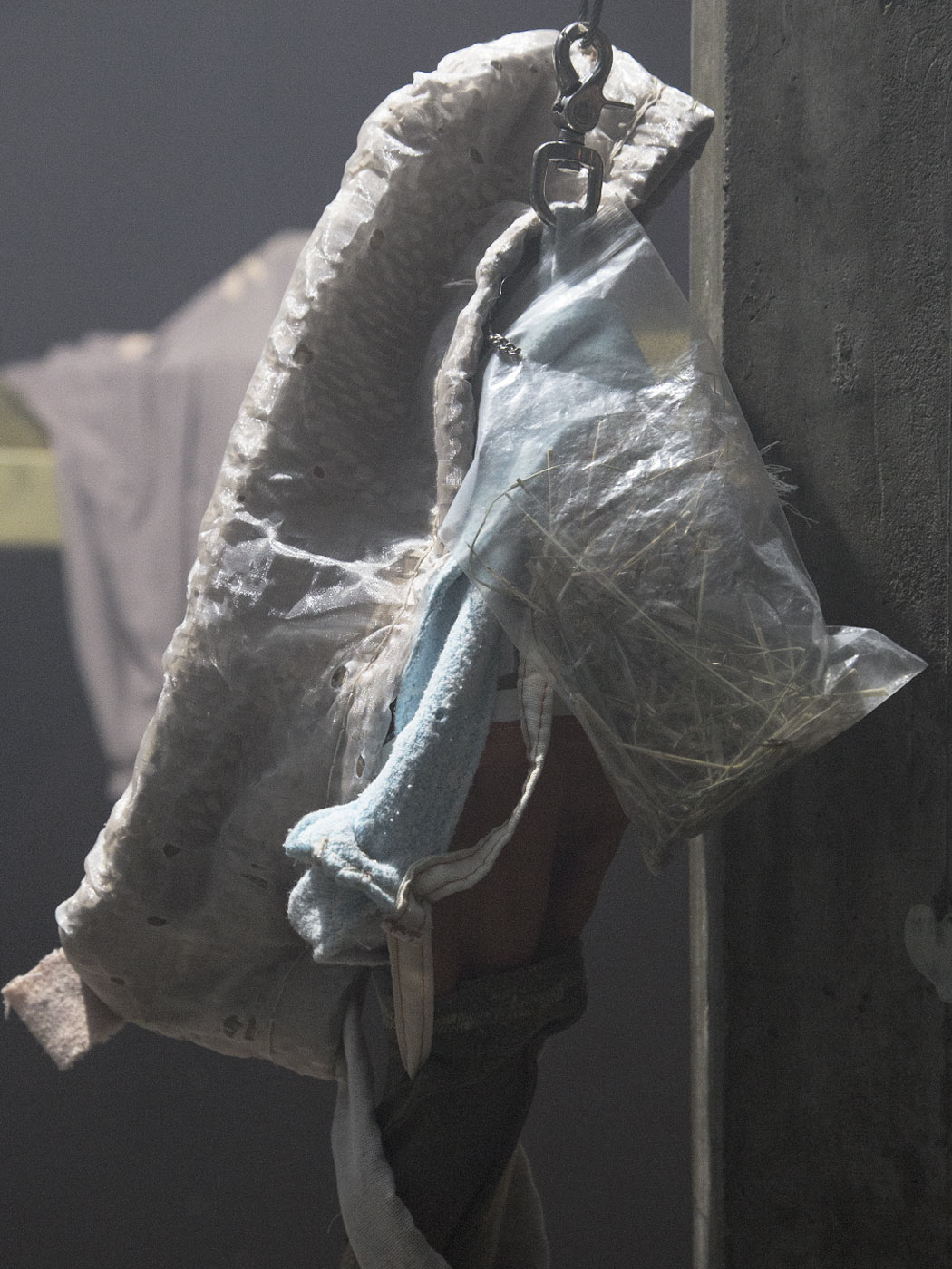 A bag filled with hay, leaning against an elongated, beige object made of laserscrim, underneath a blue pole made of textile, from which white textile strips snake out along a sleeve hem.