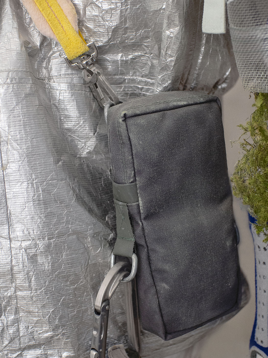 A green-grey shoulder bag with a 1:2 aspect ratio. The strap attached to the bag is rose-colored and has yellow leather ends. There are D-rings on the bag and an aluminum carabiner is attached to one of the rings.