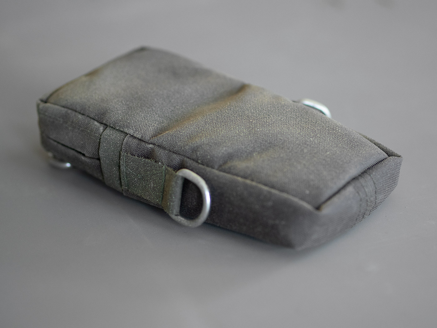 A green-grey shoulder bag with galvanized metal d-rings. The bag has an aspect ratio of 1:2.
