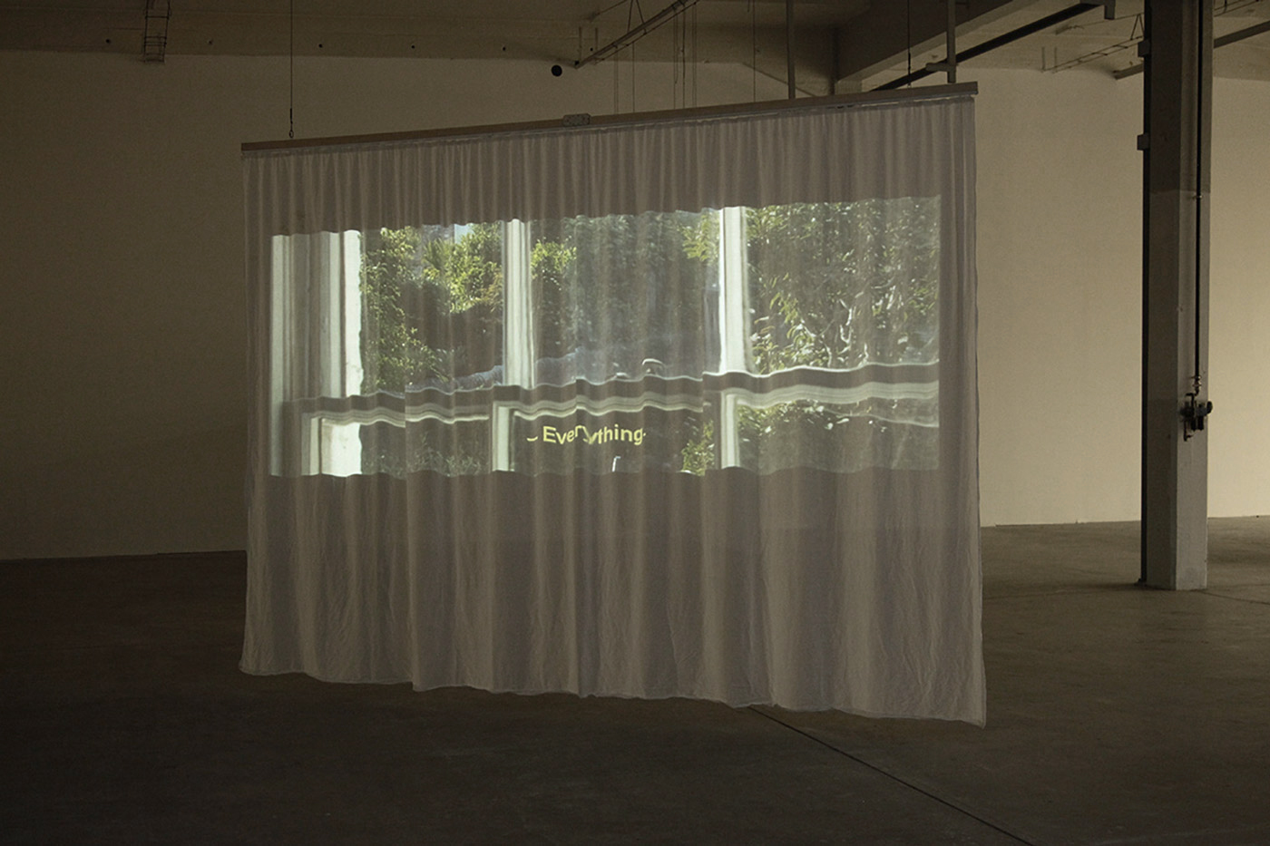 A gathered curtain hanging in an industrial space flooded with evening light, on which a video still of a view out of the window into the greenery can be seen. The subtitle in the picture reads Everything.